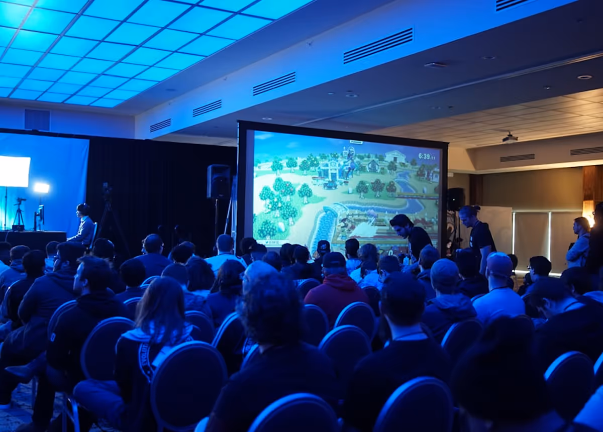 Audience seated in a dimly lit room watching a large screen displaying a video game with a vibrant blue ceiling light.