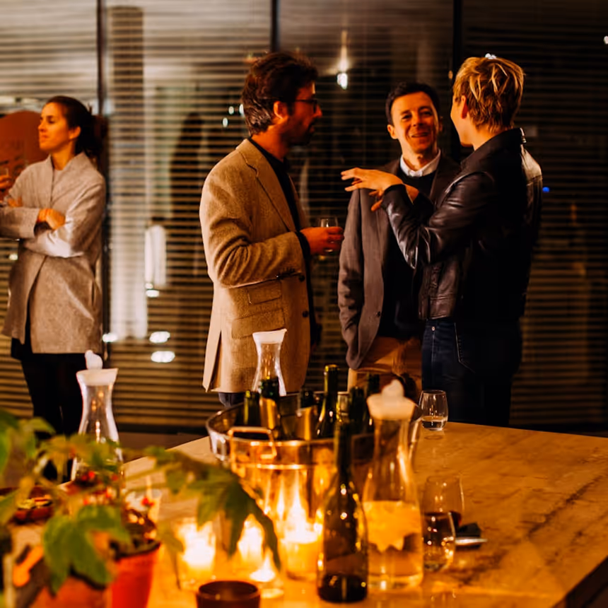 Three people engaged in conversation at a dimly lit indoor social gathering with drinks on a table in the foreground.