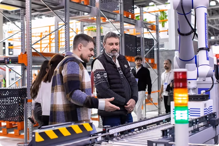Two men observing a conveyor belt and robotic arm in a modern industrial warehouse.