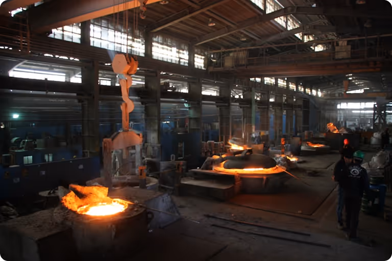 Interior of a metal foundry with molten metal glowing in large crucibles and a worker walking nearby.
