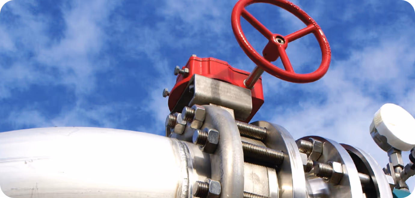 Close-up of an industrial metal pipe with a red valve handle and bolts against a blue sky with clouds.
