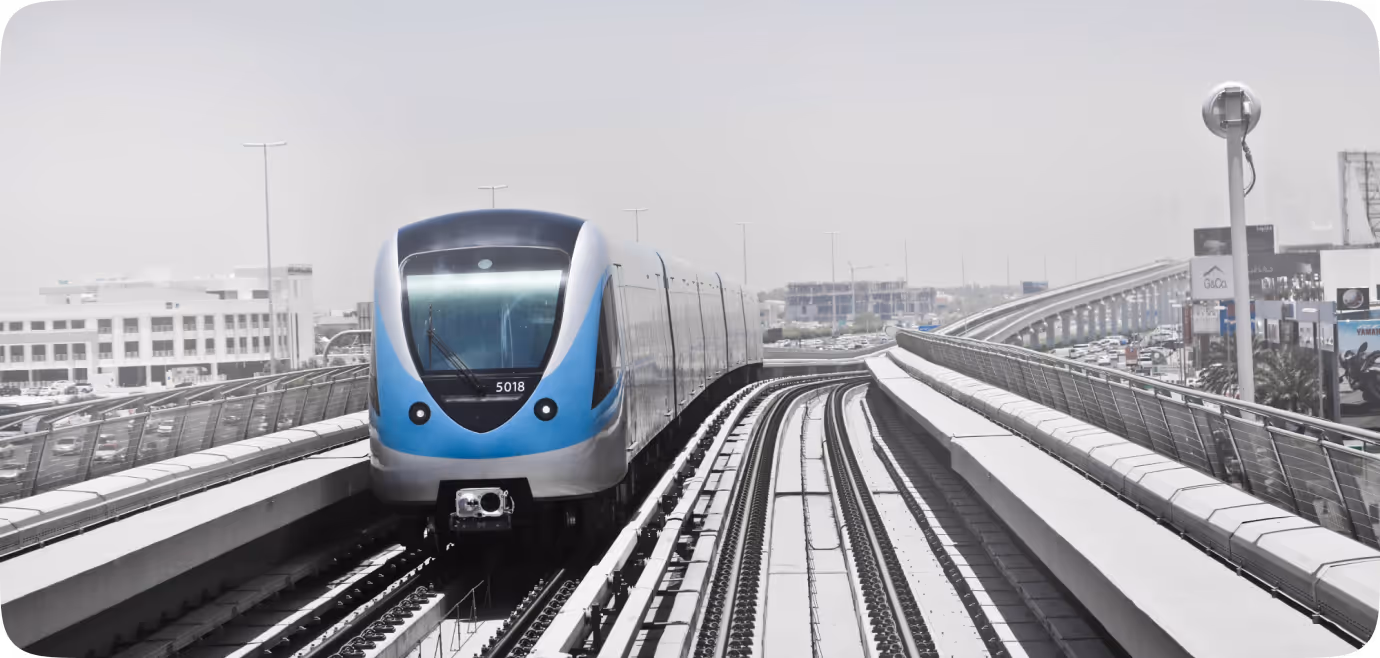 Modern blue and silver metro train traveling on elevated tracks in an urban area with buildings and traffic in the background.