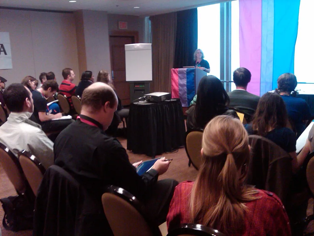 Robyn Ochs speaking to a room at the Bisexual, Fluid, Pansexual, Queer-identified and Allies Organizing Institute, 2010, with a bisexual pride flag behind her
