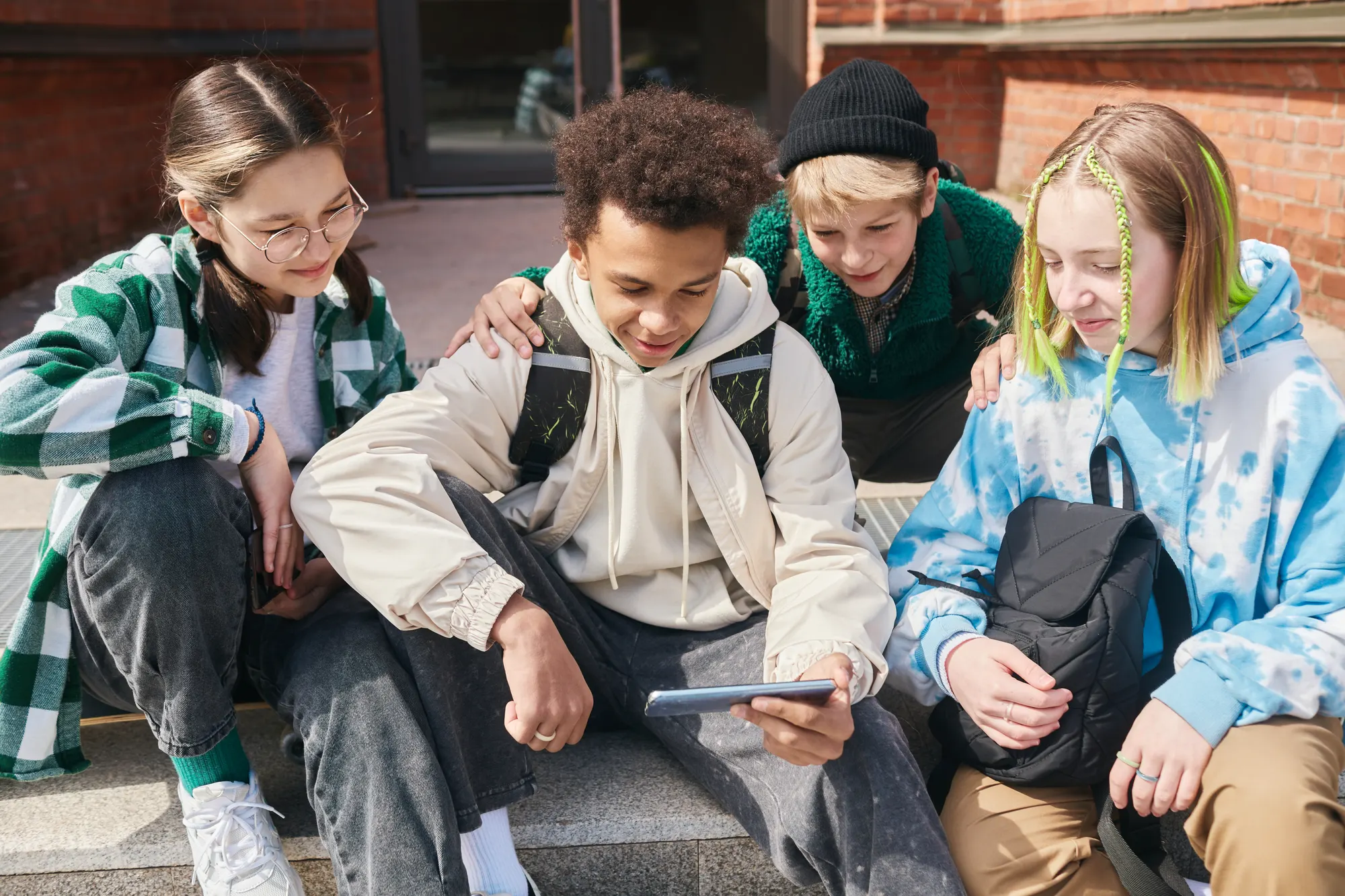 Four teenagers with backpacks sitting together on the steps outside a brick school building, looking at a phone and smiling
