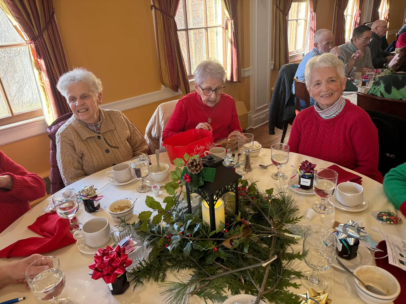 Three smiling guests in holiday sweaters at a round table with a lantern and evergreen centerpiece