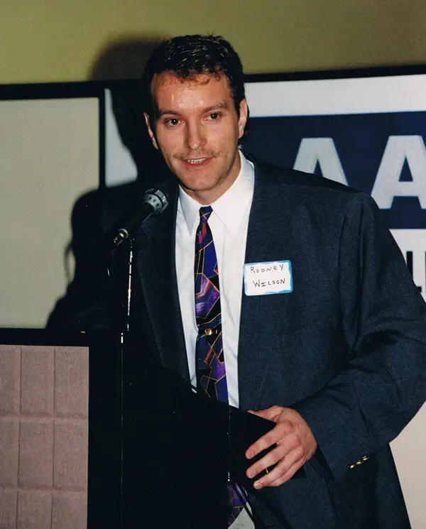 Rodney Wilson, in a dark suit and patterned tie with a handwritten name sticker, speaking at a microphone at a podium