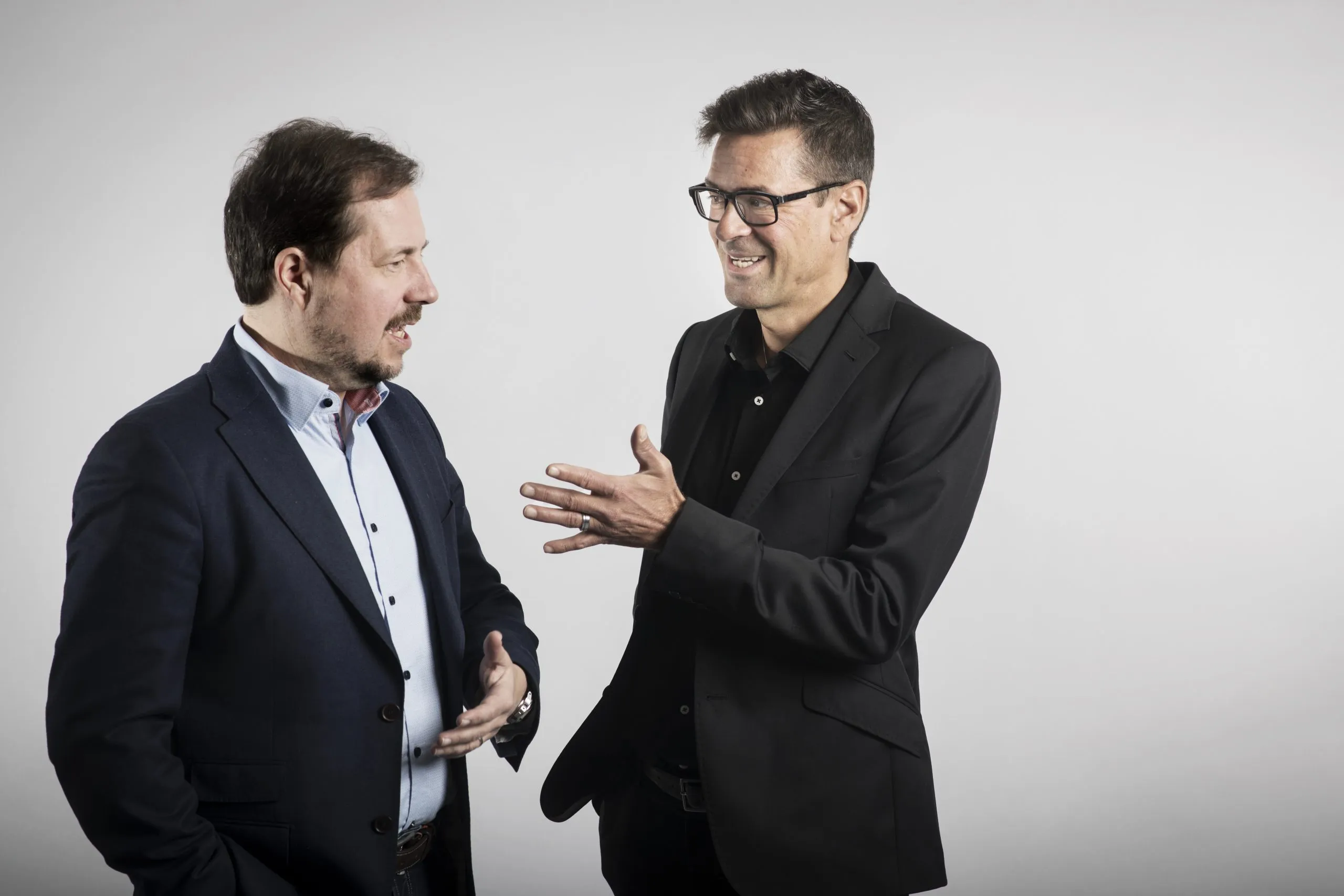 Kaksi miestä seisoo ja keskustelee vaaleanharmaalla taustalla. Vasemmalla oleva mies, jolla on parta ja viikset, pukeutuu tummansiniseen pikkutakkiin ja vaaleansiniseen paitaan. Hän katsoo oikealla olevaa miestä. Oikealla oleva mies, jolla on silmälasit ja musta pikkutakki mustan paidan päällä, hymyilee ja elehtii oikealla kädellään. Molemmat miehet ovat iloisia ja katsovat toisiaan.