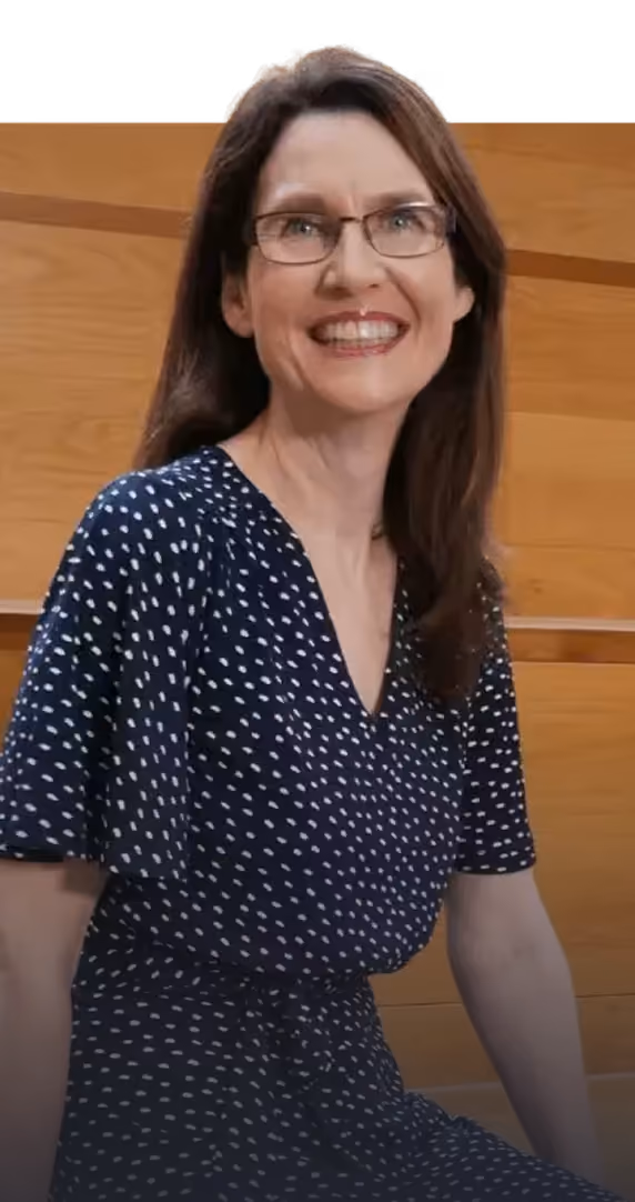 Smiling woman with glasses wearing a navy blue dress with white spots sitting against a wooden background.