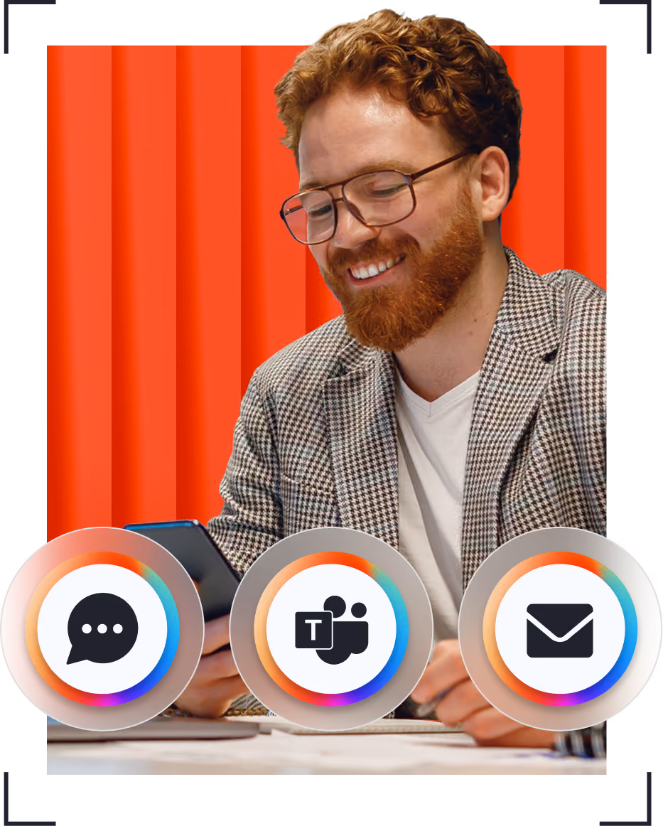 Smiling man with glasses and red hair using phone at desk with chat, Teams, and email icons.