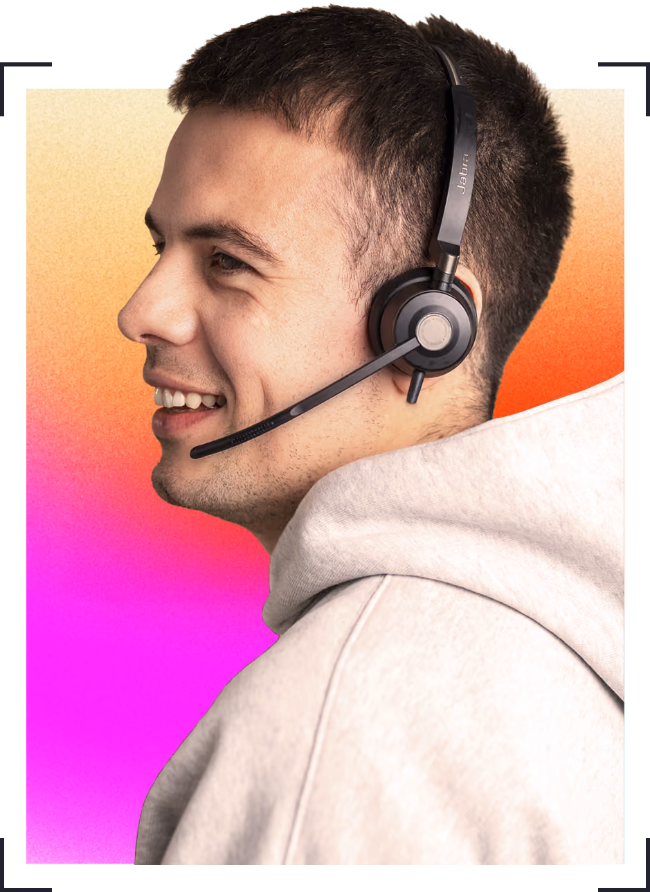 Young man smiling while wearing a headset with microphone and a light hoodie against a pink-orange background.