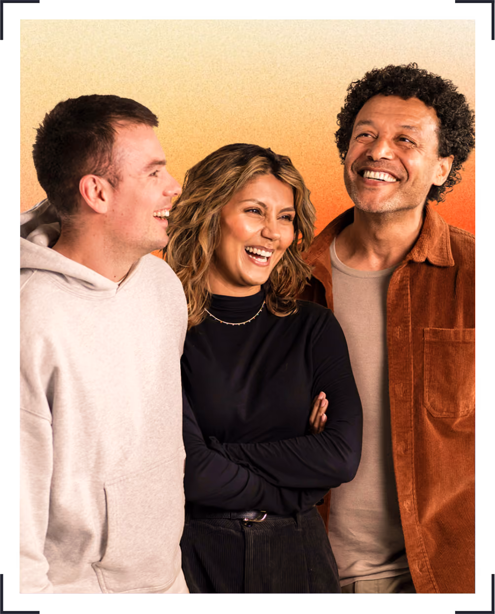 Three adults smiling together, one in beige hoodie, one in black top, and one in brown jacket.