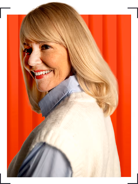 Smiling older woman with shoulder-length blonde hair in white sweater and blue shirt on orange background.