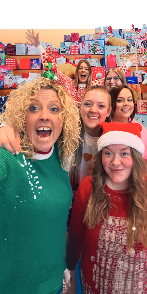Group of six smiling women in festive Christmas sweaters and Santa hat with wrapped gifts behind.