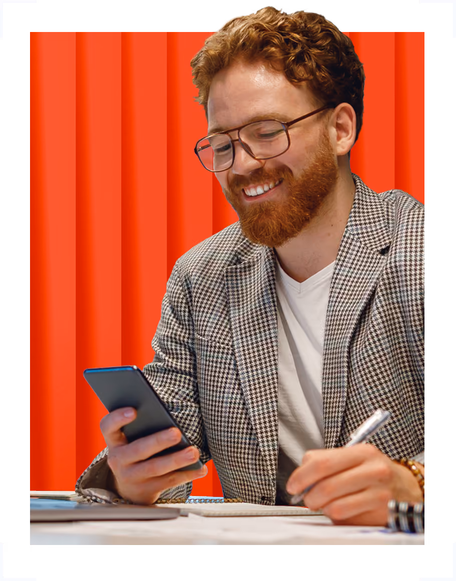 Smiling man with glasses and red hair using phone at desk with chat, Teams, and email icons.