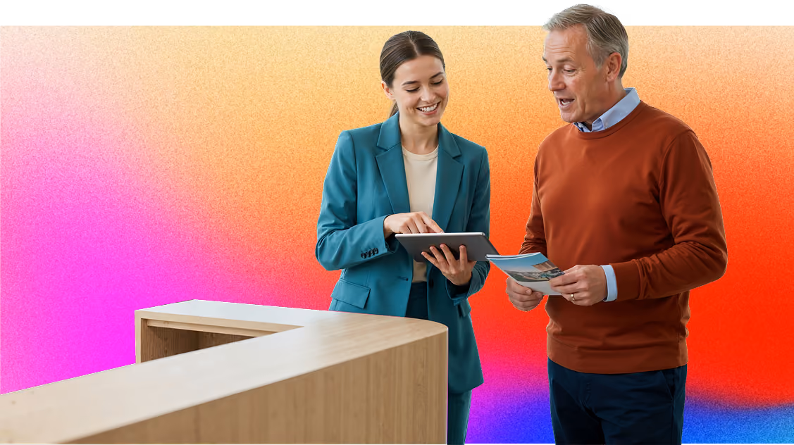 A woman in a teal suit shows something on a tablet to a man in a brown sweater who holds a brochure, standing by a wooden reception desk against a colourful gradient background.