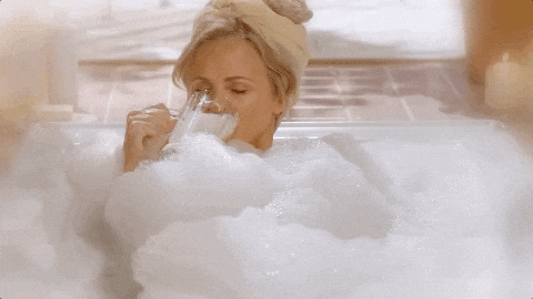 Woman relaxing in a bubble bath drinking a glass of milk or white beverage, surrounded by fluffy bubbles.