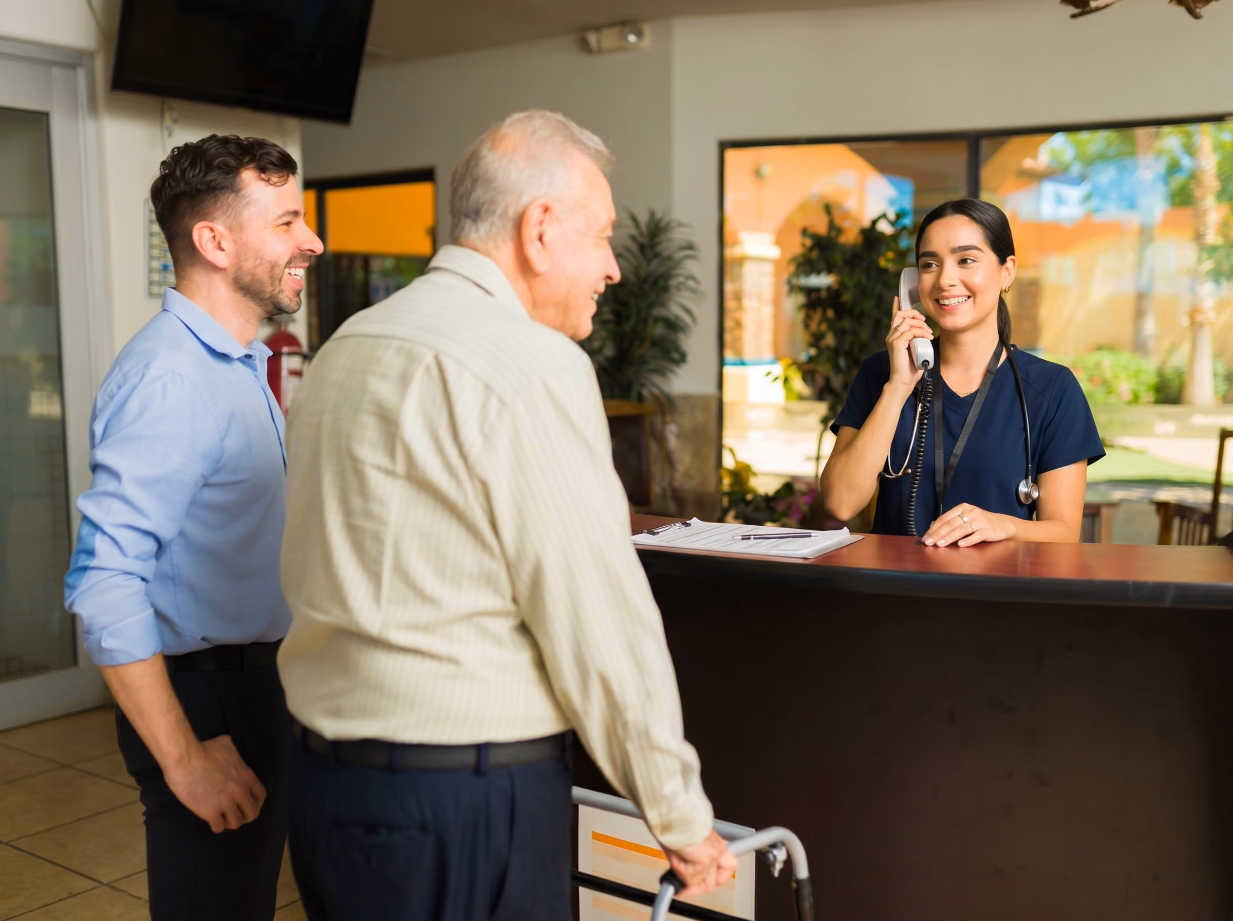 Nurse at reception desk talking on phone while elderly man with walker and younger man stand nearby.