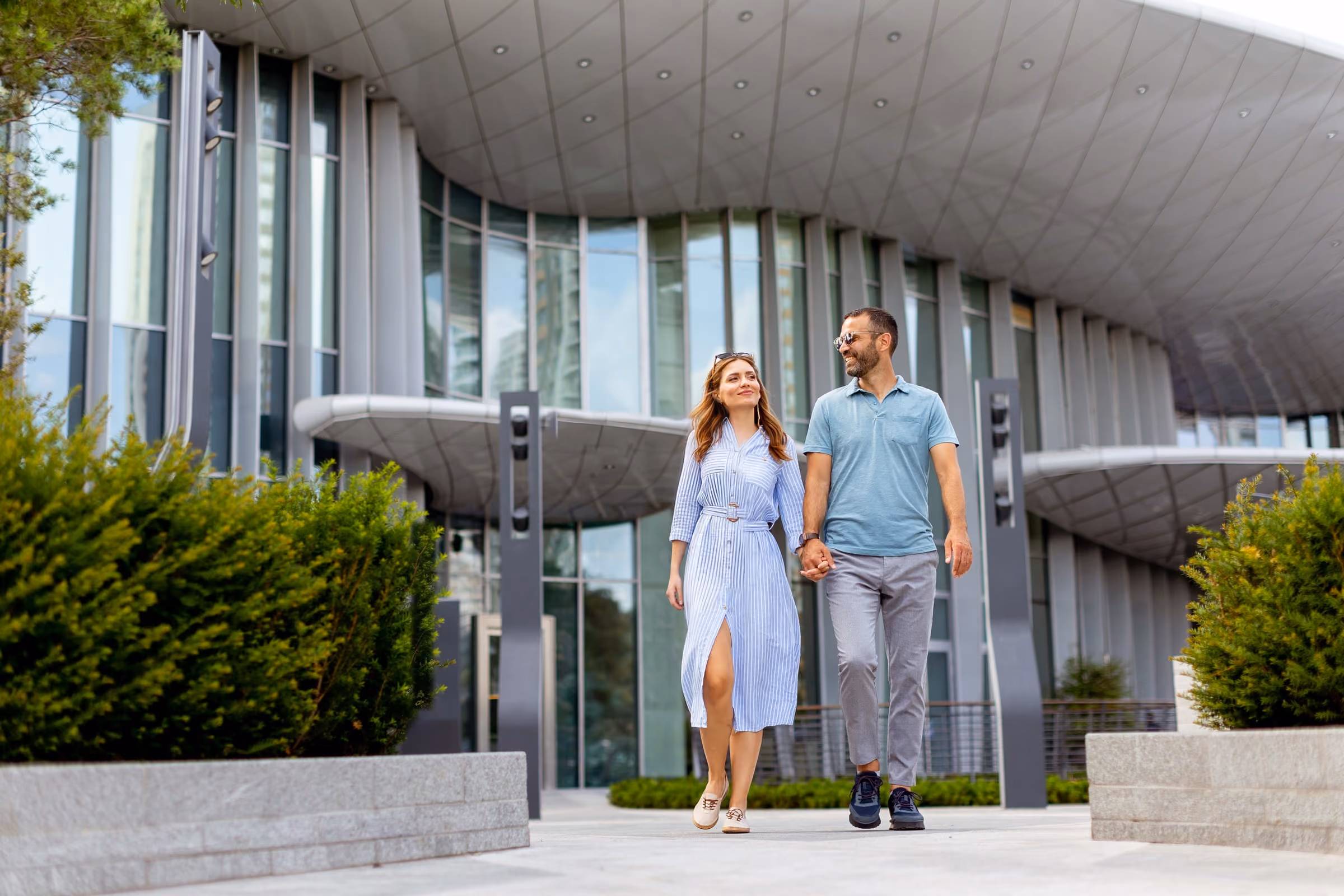 Man and woman holding hands while walking past modern glass building with greenery.