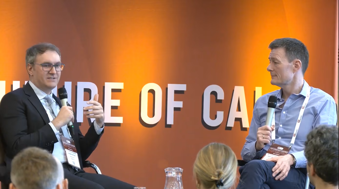 Two men in a discussion seated on stage holding microphones with audience heads visible in foreground.