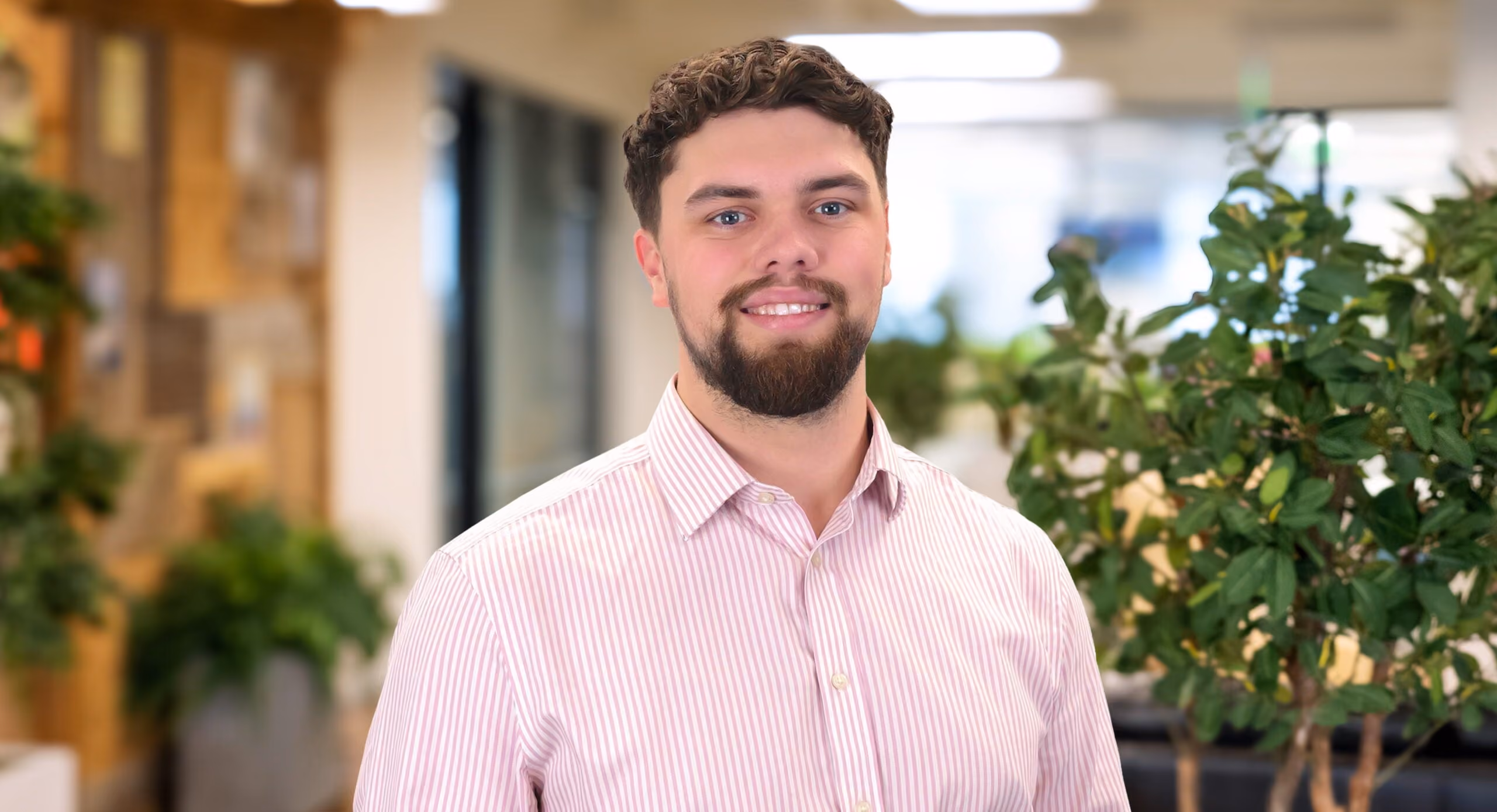 Smiling man with short curly hair and beard wearing a striped button-up shirt indoors by plants.