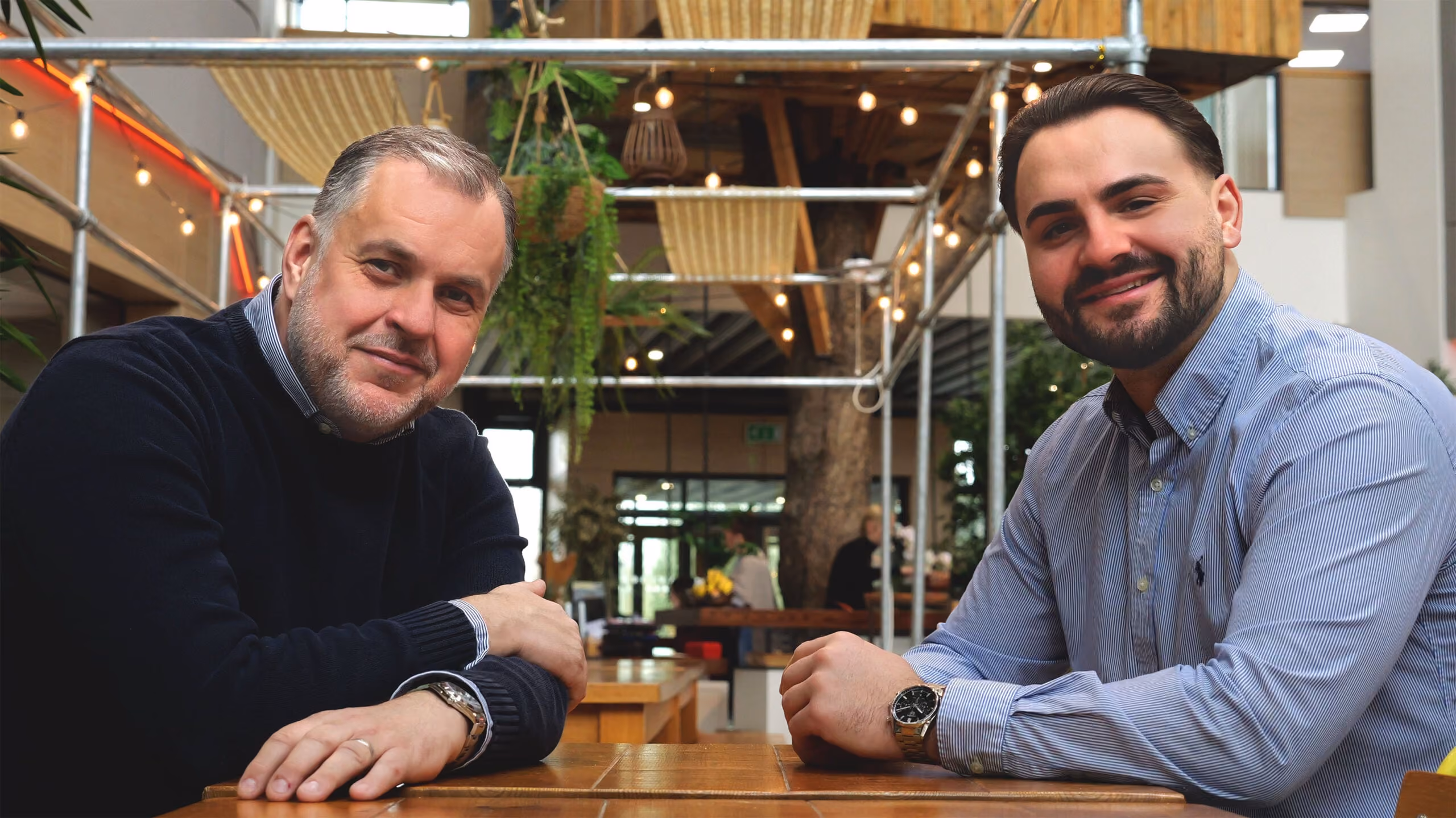 Two men sitting at a wooden table, smiling toward the camera in a modern, plant-filled indoor space.