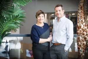 A woman with short brown hair wearing a navy and black top stands beside a man in a light grey shirt with his hands in his pockets, both smiling at the camera in an indoor setting with plants and furniture around.