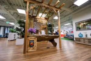 Indoor showroom featuring a wooden pergola with climbing plants above a rustic wooden table, surrounded by furniture and decorative items on a wooden floor.