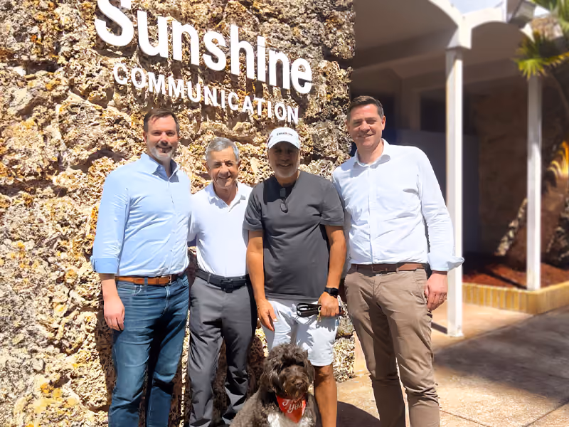 Four men standing in front of a Sunshine Communication sign with a seated brown dog wearing a red bandana.