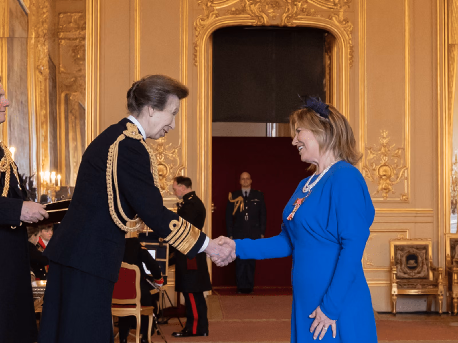 Two women shaking hands in an ornate room, one in a navy uniform, the other wearing blue with medals.
