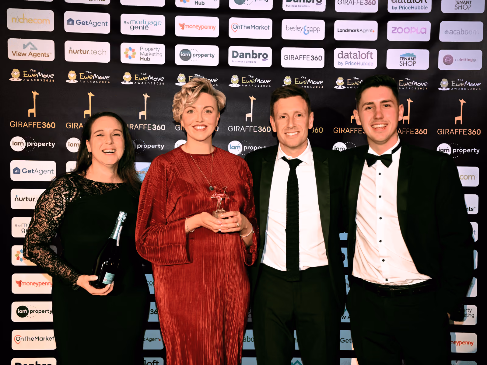 Four people in formal attire posing in front of event backdrop holding an award and a bottle of champagne.