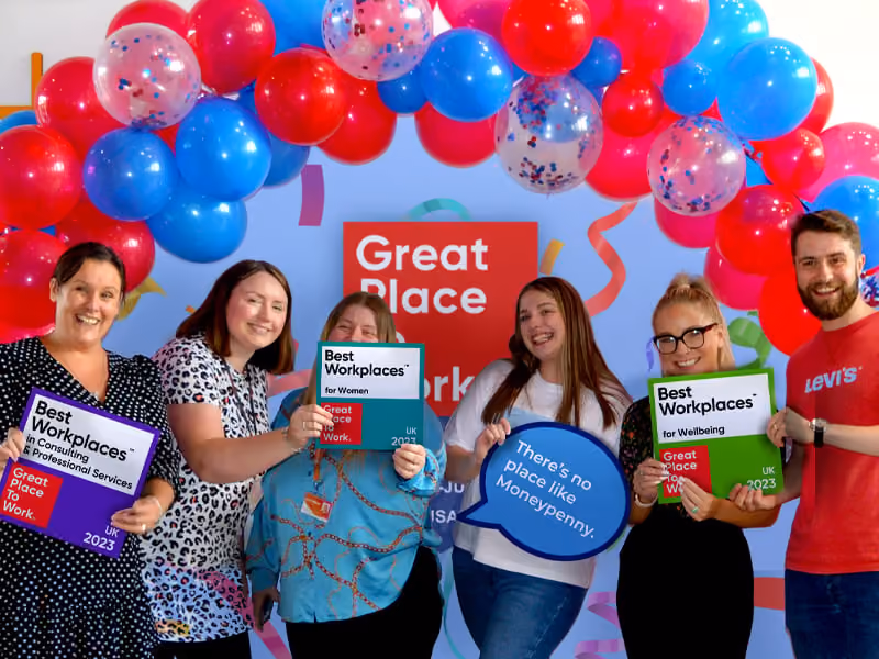Six smiling people holding Best Workplaces award plaques and a speech bubble under a red and blue balloon arch.