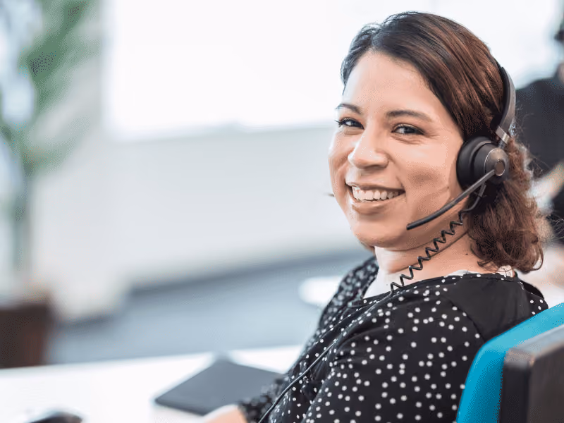 Smiling woman wearing a headset, working in a bright office environment.