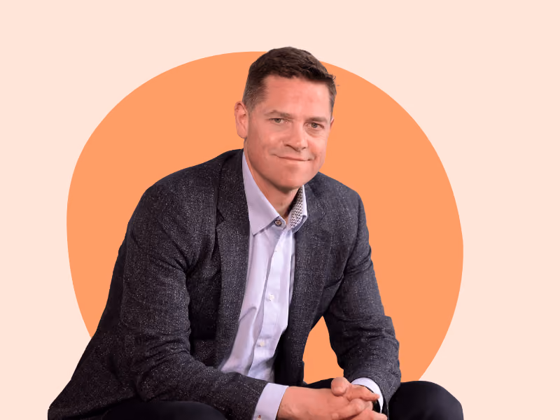Man in dark blazer and light shirt sitting against a pastel peach and orange circular background.