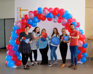 Five smiling people stand under an arch made of red, blue, and white balloons, holding signs with positive messages about great workplaces and company culture.