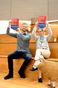 Two people sitting on a wooden bench holding red and blue signs that read 'Great Place to Work Certified', with a large sheep sculpture in front of them.
