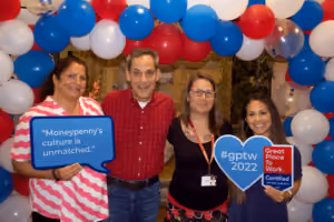 Four people smiling under a red, white, and blue balloon arch holding Great Place to Work signs.