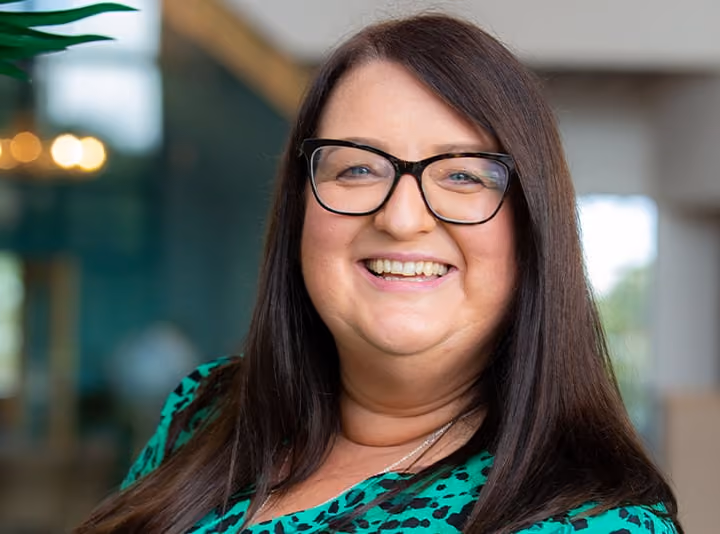 Smiling woman with long dark hair and glasses wearing a green and black patterned top.