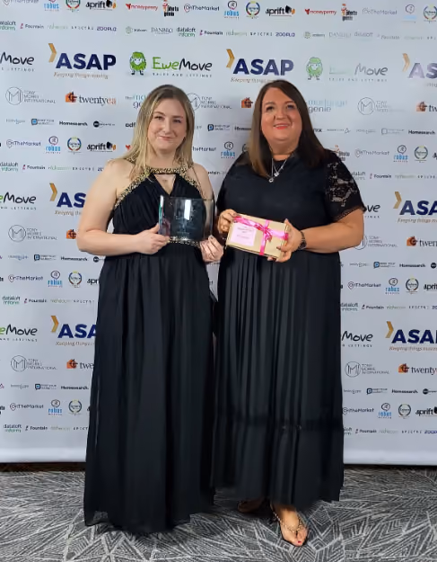 Two women in black dresses holding awards and standing in front of a sponsor banner at an event.