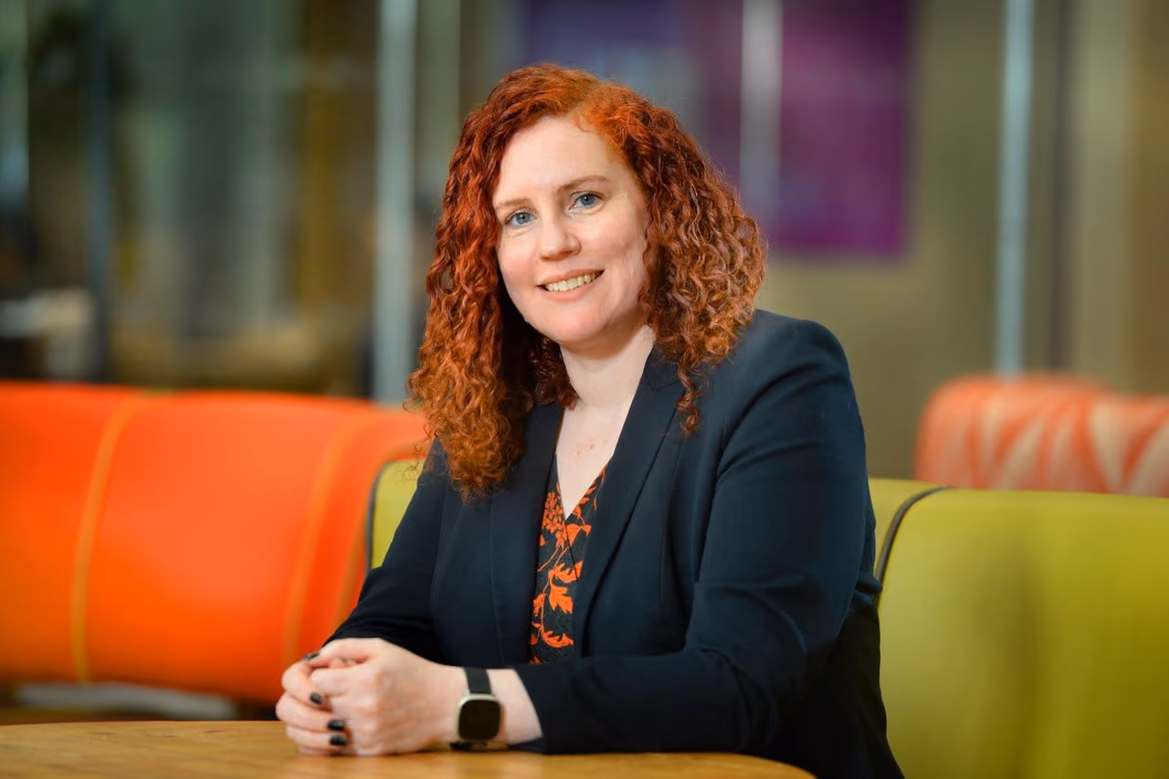 Smiling woman with curly red hair sitting at a table wearing a black blazer and smartwatch.