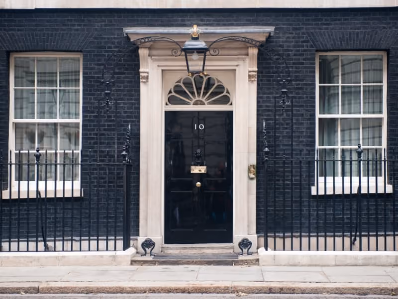 Black front door of 10 Downing Street, flanked by two windows and black iron fences.