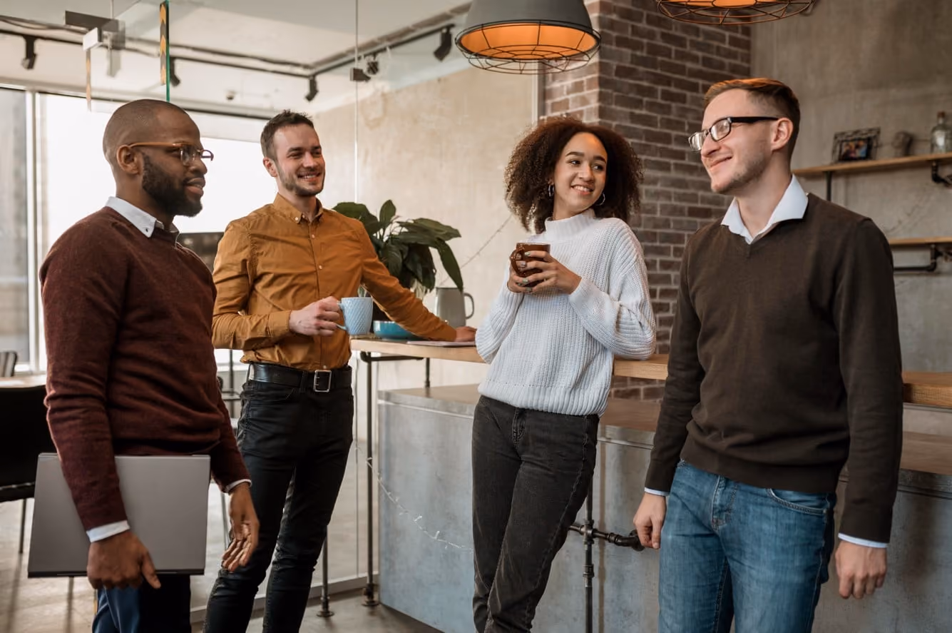 Four young coworkers chatting and smiling in a modern office kitchen area with coffee mugs.