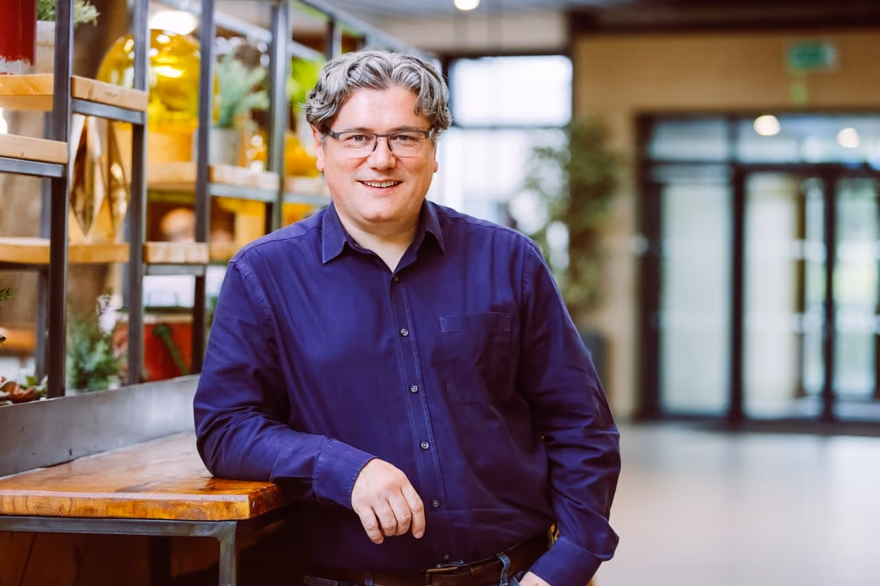Man with glasses and gray hair in a purple shirt leaning on a wooden table in a modern indoor space.