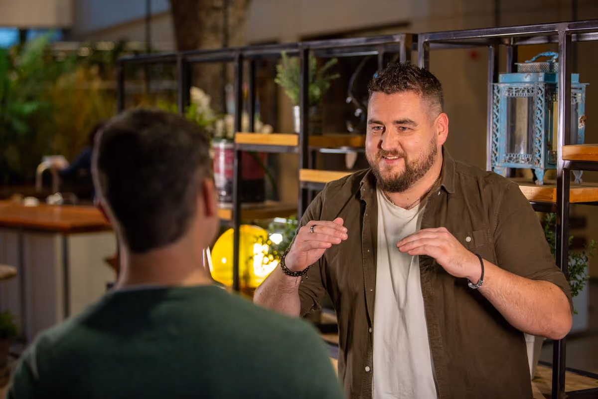 Smiling man with beard gesturing while talking to another man in a cozy, decorated indoor space.