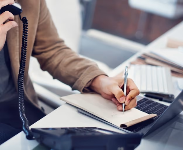 Person on phone writing notes on a notepad at a desk with a laptop and keyboard.