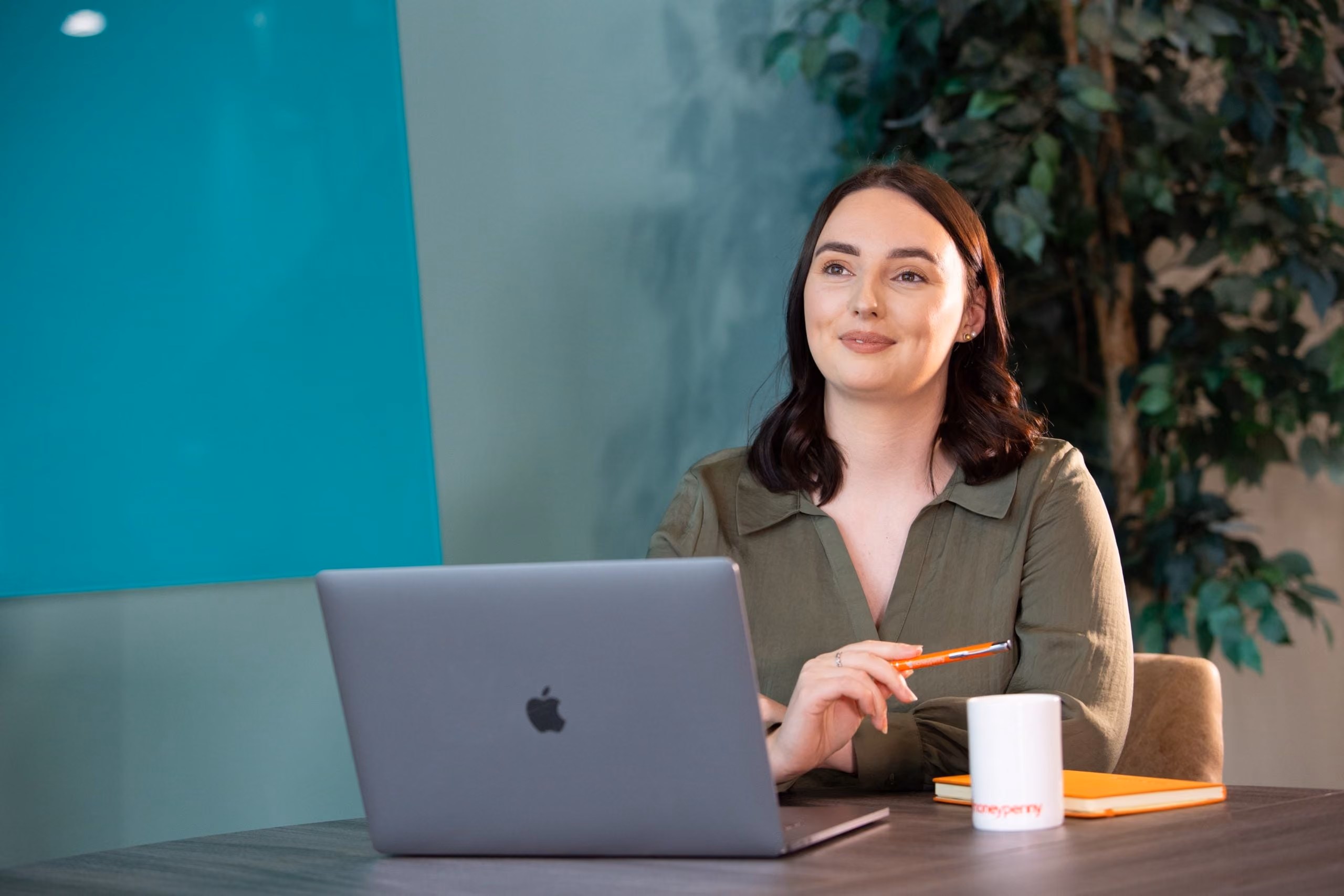 Woman with dark hair sitting at a desk, holding an orange pen, with a laptop, notebook, and mug.