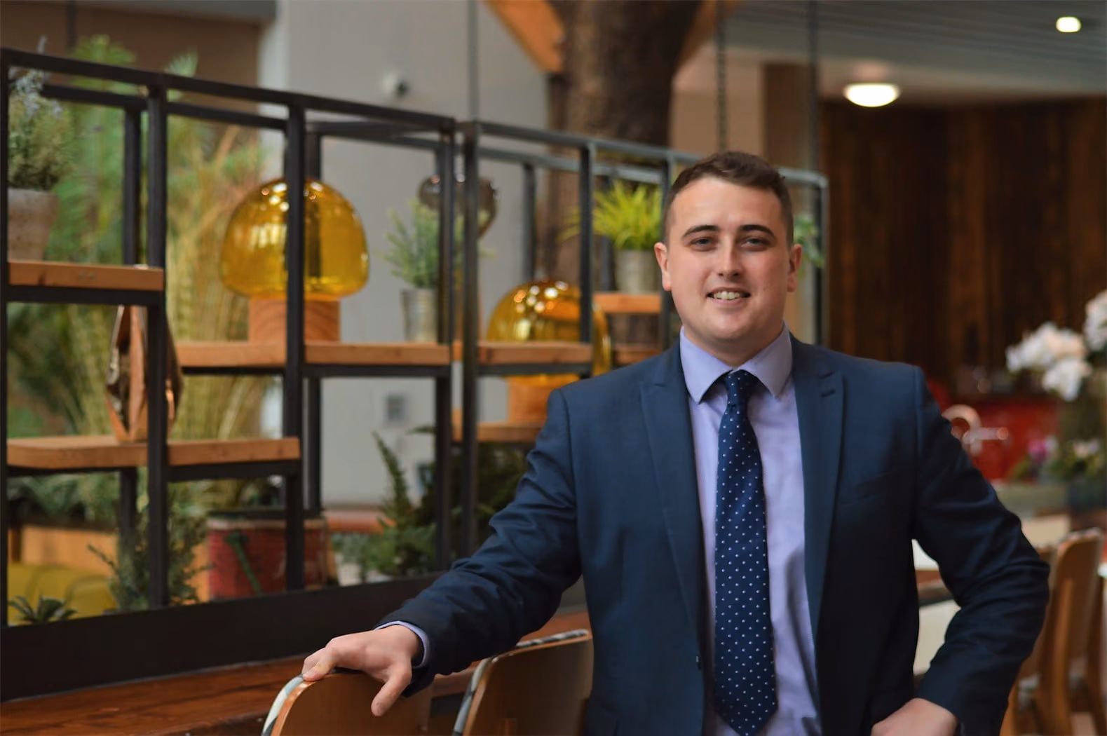 Smiling man in suit and polka dot tie sits in modern indoor space with plants and amber lights.
