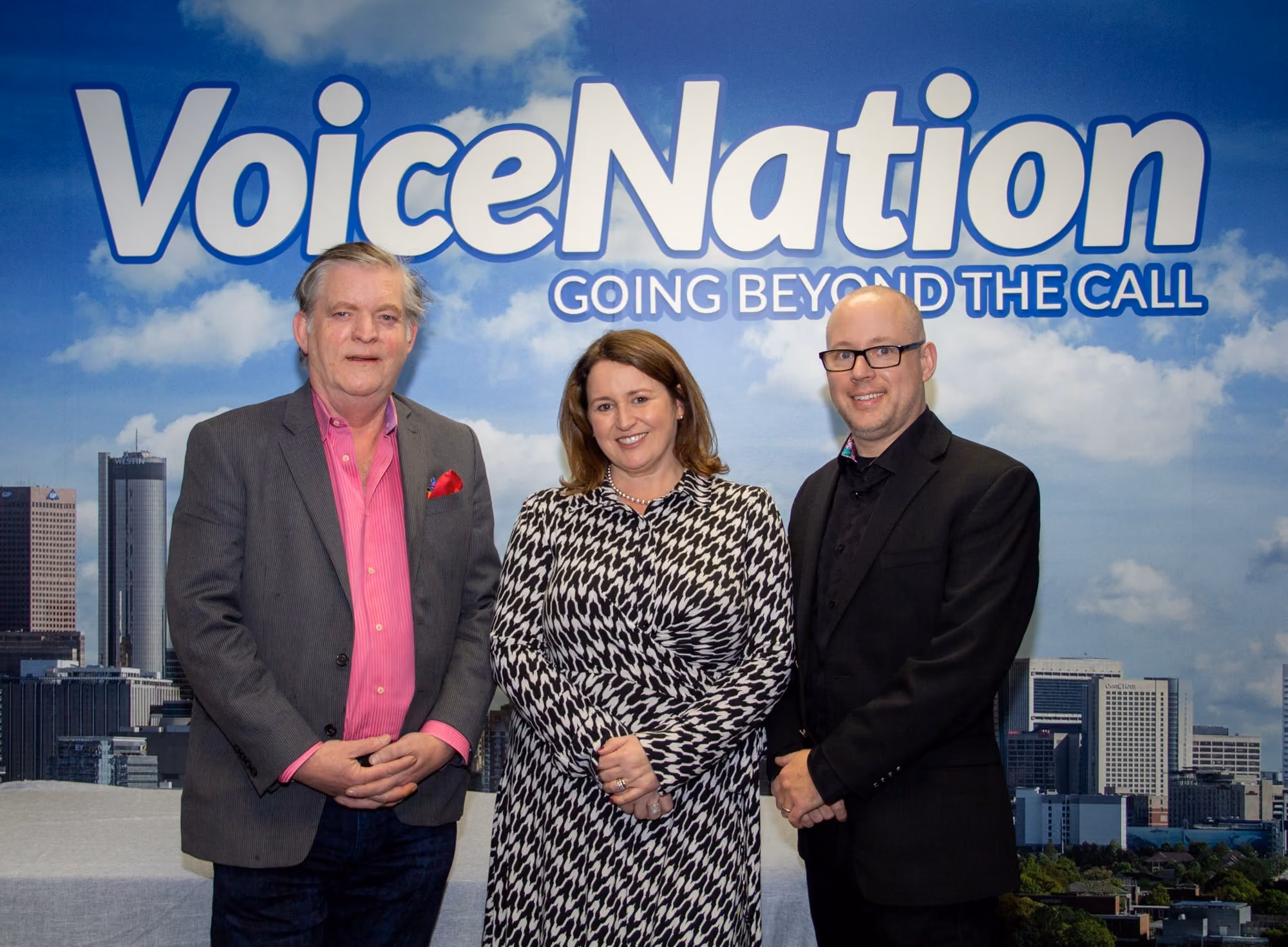 Three people posing in front of a VoiceNation sign with cityscape and cloudy sky background.