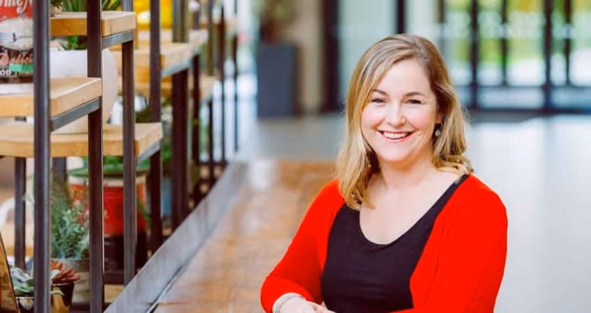 Smiling woman with blonde hair wearing red cardigan and black top sitting indoors near wooden shelves.