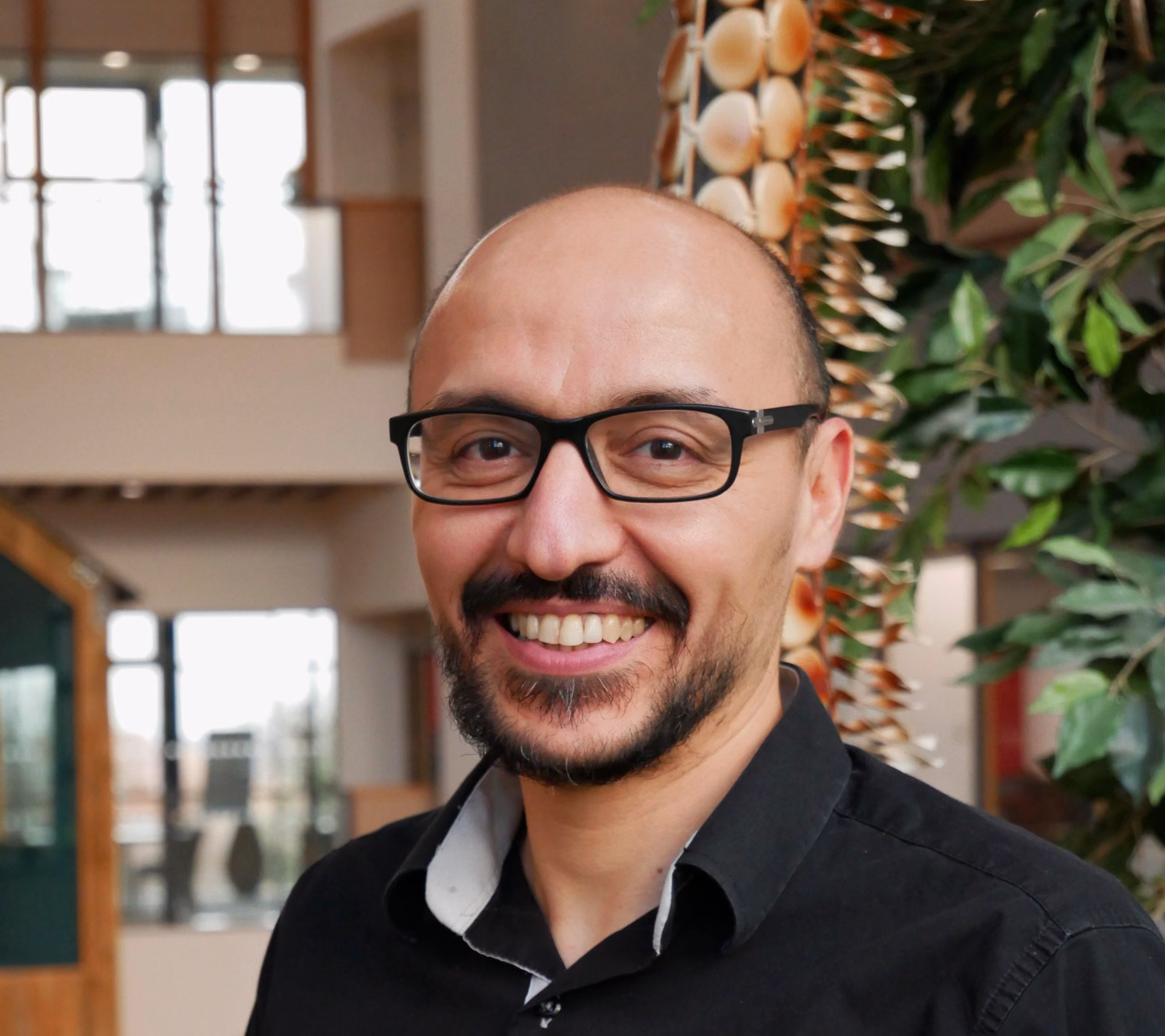 Smiling man with glasses and a beard wearing a black shirt indoors with plants in the background.