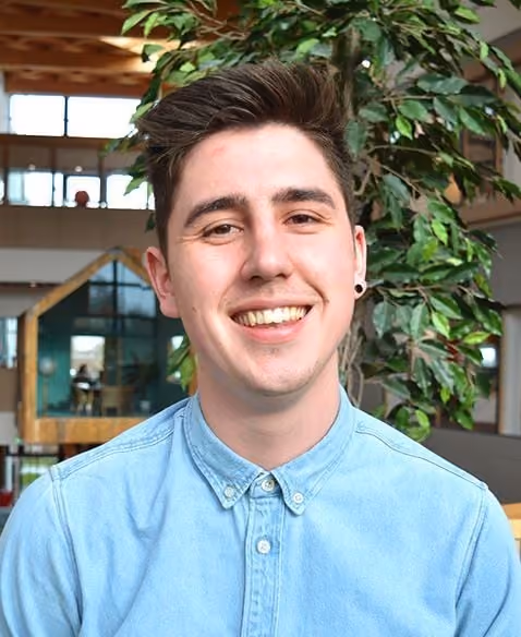 Young man smiling in a light blue button-up shirt with a plant and indoor office background.